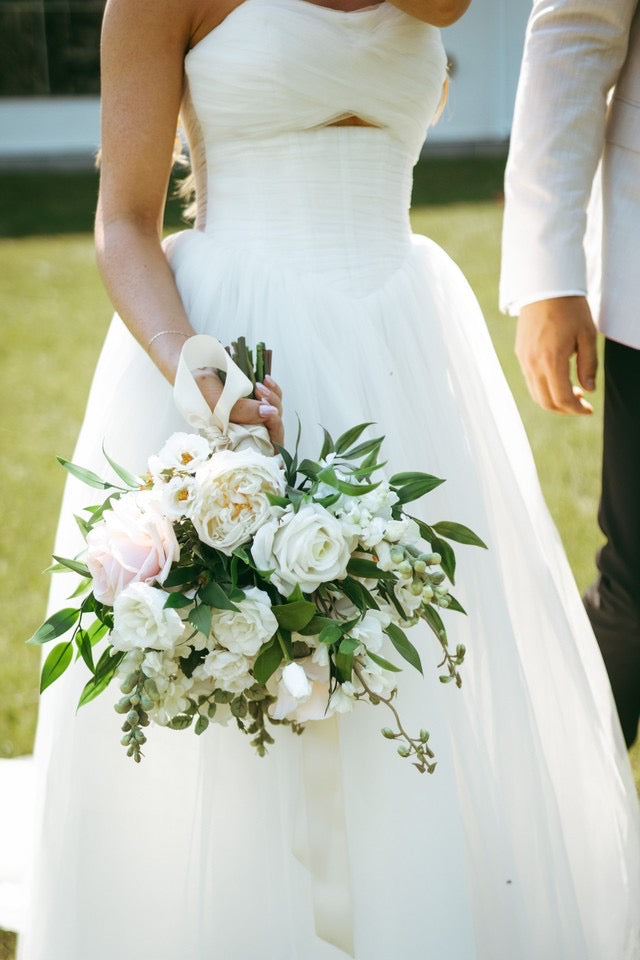 A close-up of a bride in a strapless white wedding gown holding a bouquet of white and blush pink flowers with green foliage, standing outdoors next to a groom in a white suit.