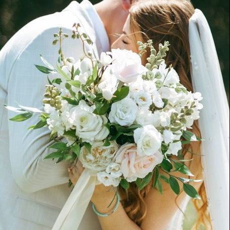 A bride in a white wedding gown and veil holding a bouquet of white and blush flowers, kissing her groom in an outdoor grassy setting.