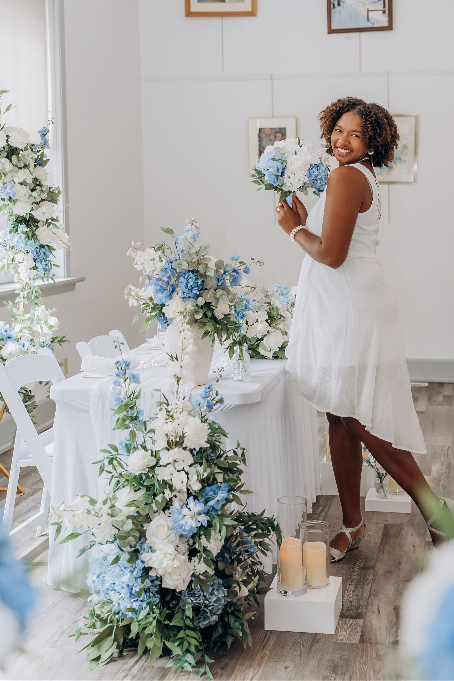 Woman in a white dress holding flowers in a room with floral arrangements and candles.